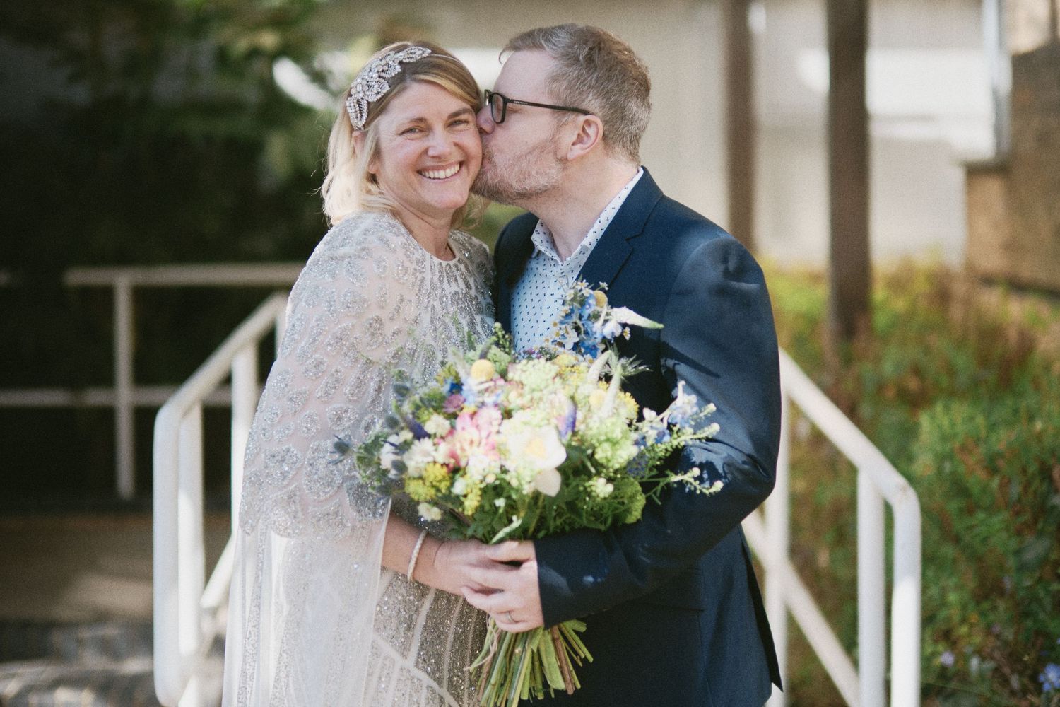 A romantic wedding moment captured in front of a white building with greenery.