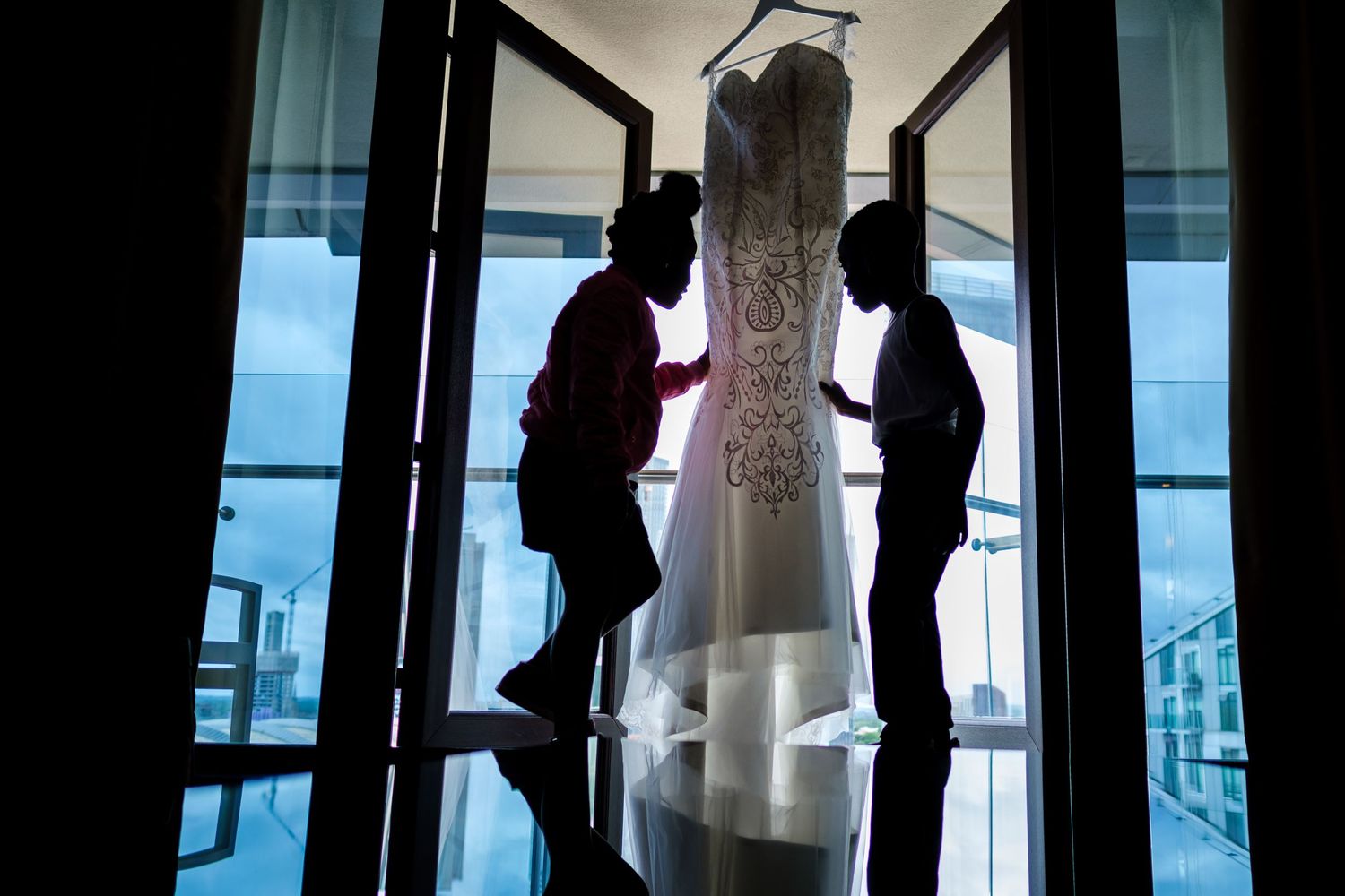 Two figures in silhouette examining a bridal gown hanging in a bright doorway.