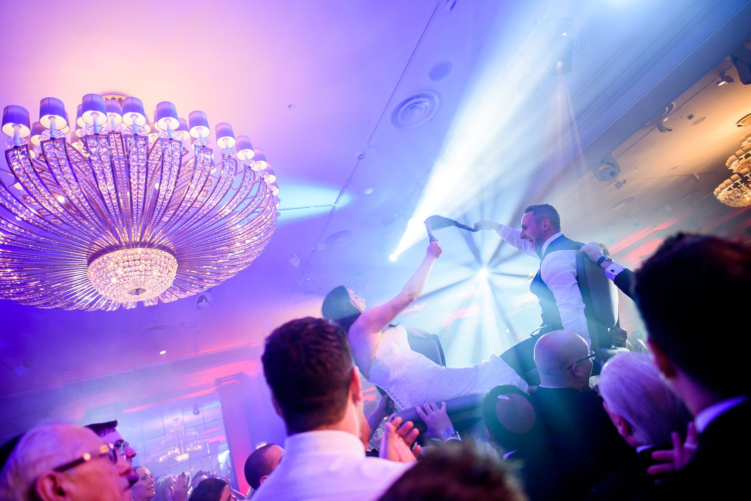 Crowd dancing under a dramatic purple-lit chandelier at an energetic wedding reception.