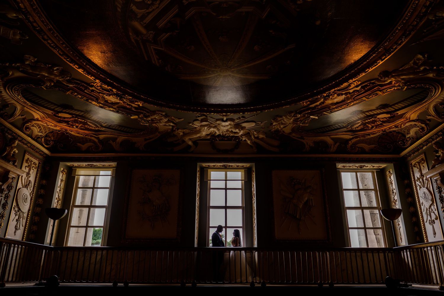 Silhouette of a couple against bright windows in a historic building with dark ceiling.