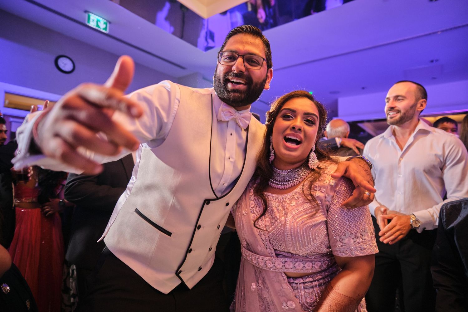 Wedding party guests laughing and dancing together on a purple-lit dance floor during a celebration.