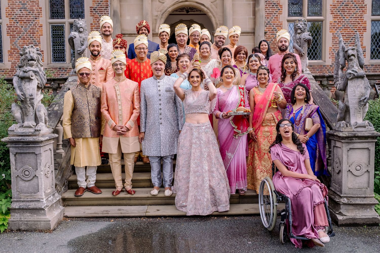 Large wedding party in colorful traditional Indian attire poses on stone steps of historic building with decorative statues.