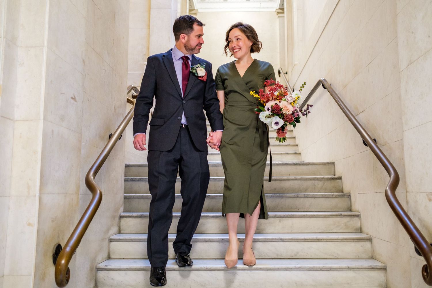A couple laughs together while walking down an elegant marble staircase in formal attire.