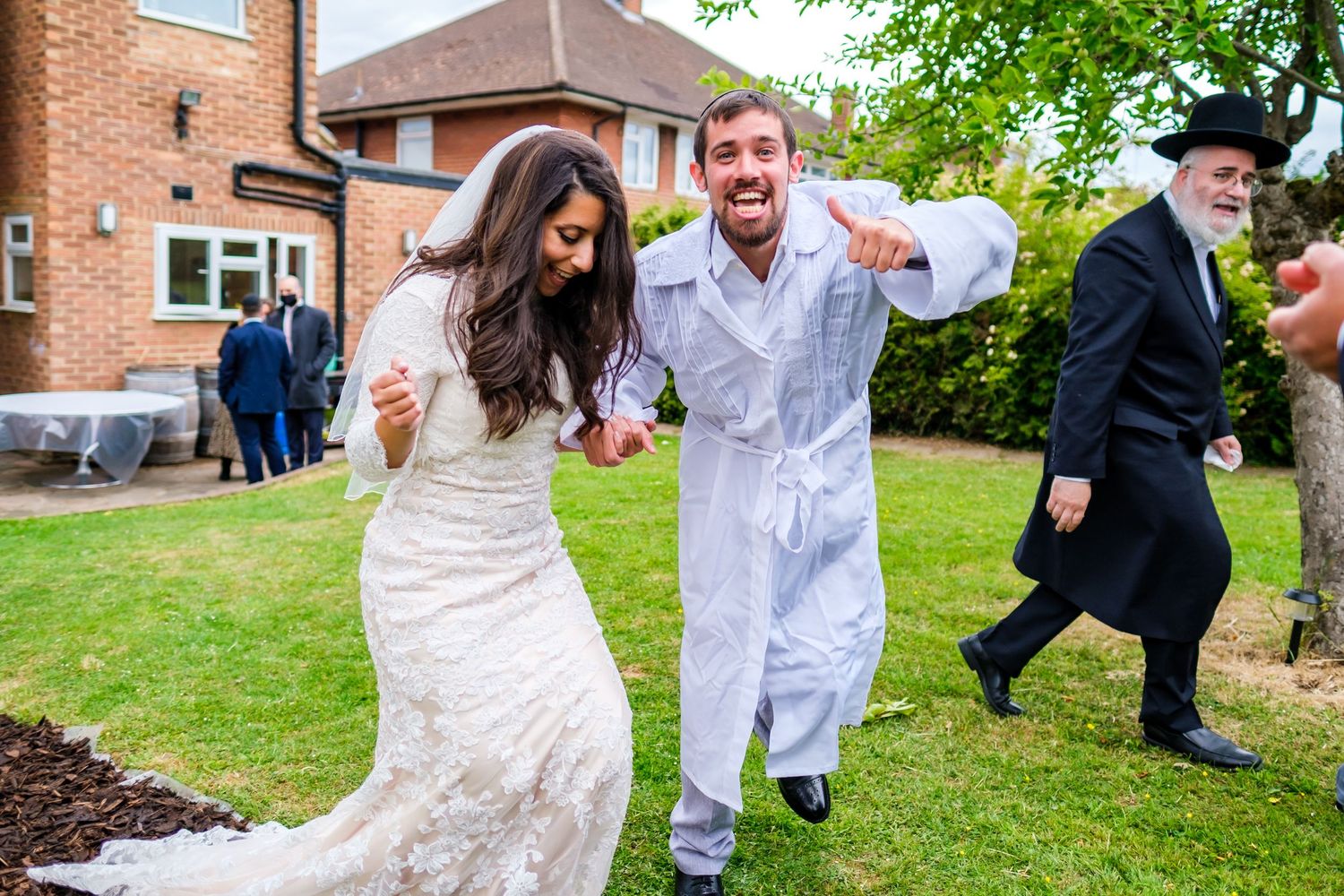 Wedding party members running and jumping playfully on a grassy lawn outside a brick building.