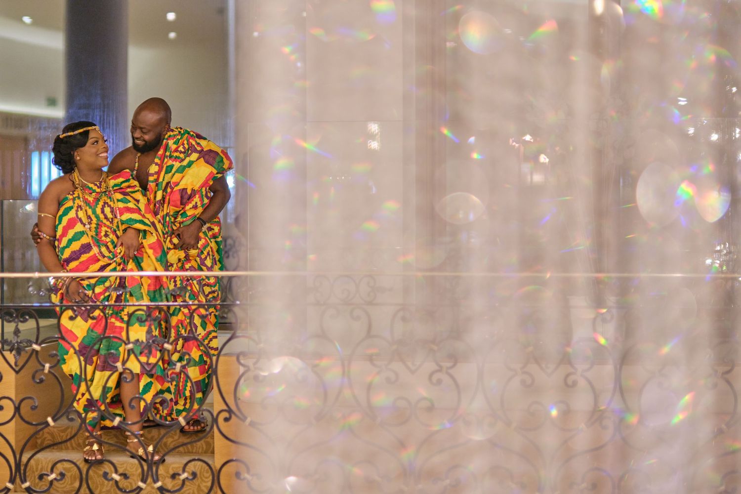 Two people wearing vibrant African print outfits stand together against a light curtain background with sparkly effects.