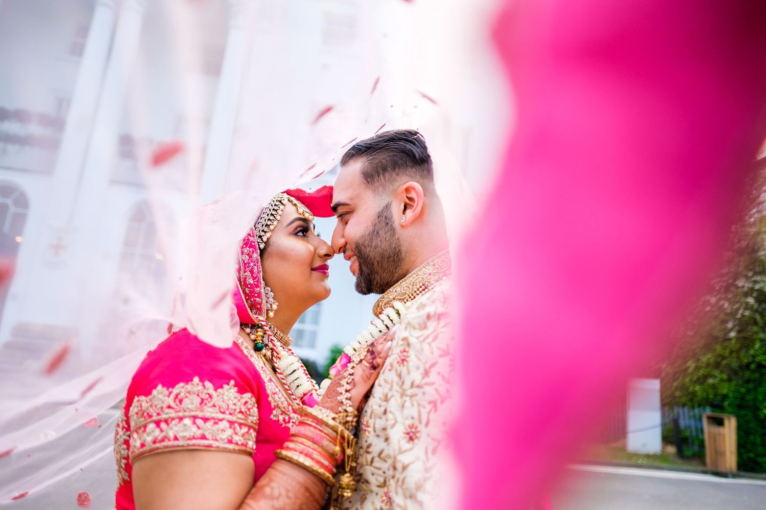 Wedding couple poses against pink backdrop in traditional South Asian bridal wear.