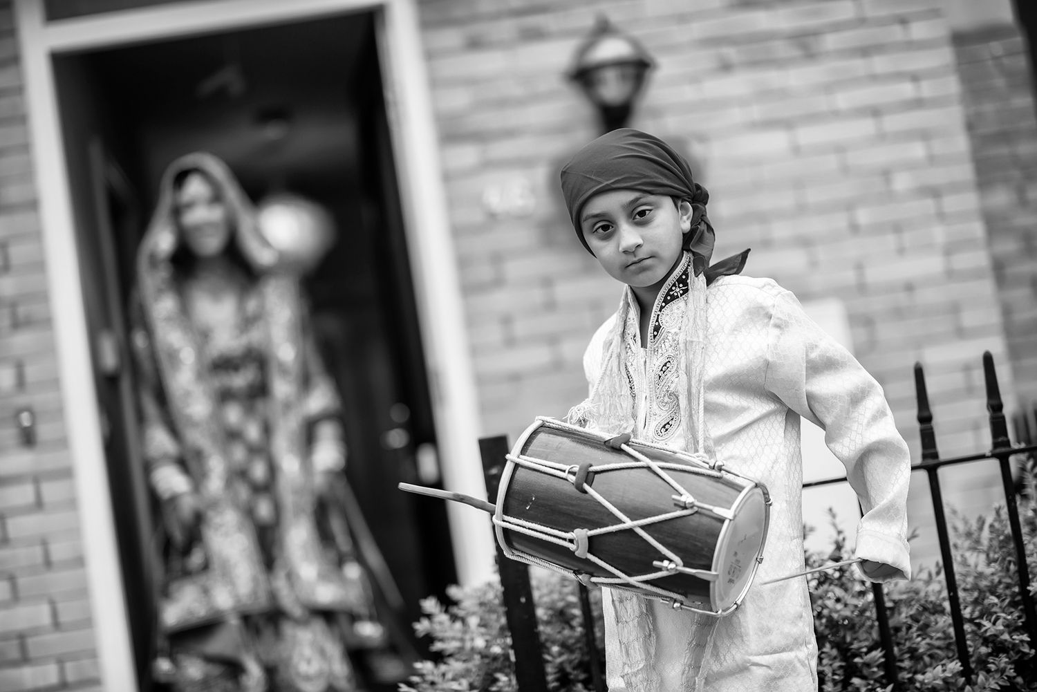 A young dhol drummer in white traditional dress performs at a celebration in black and white.