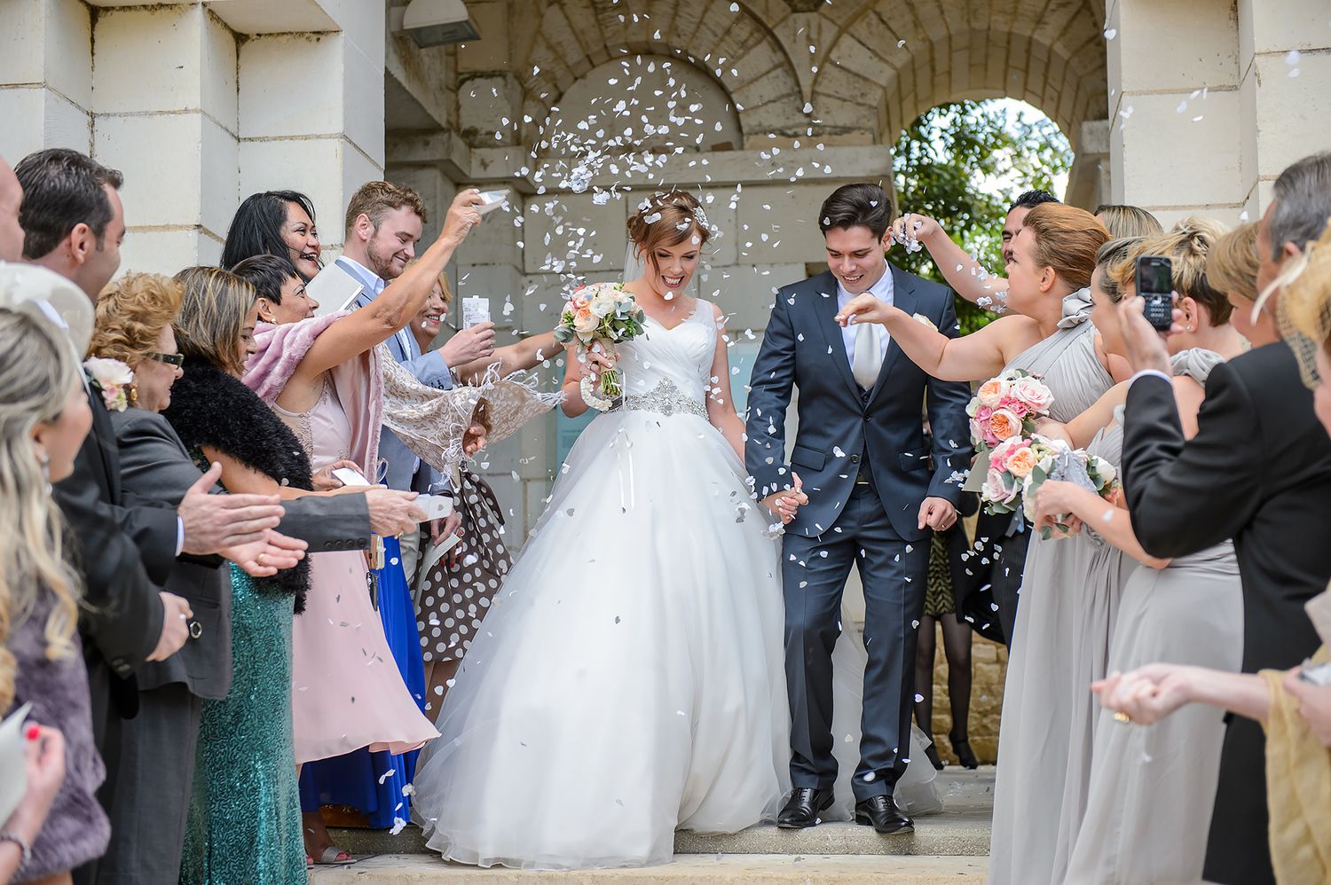 Wedding guests throwing confetti at newlyweds in front of stone archway.