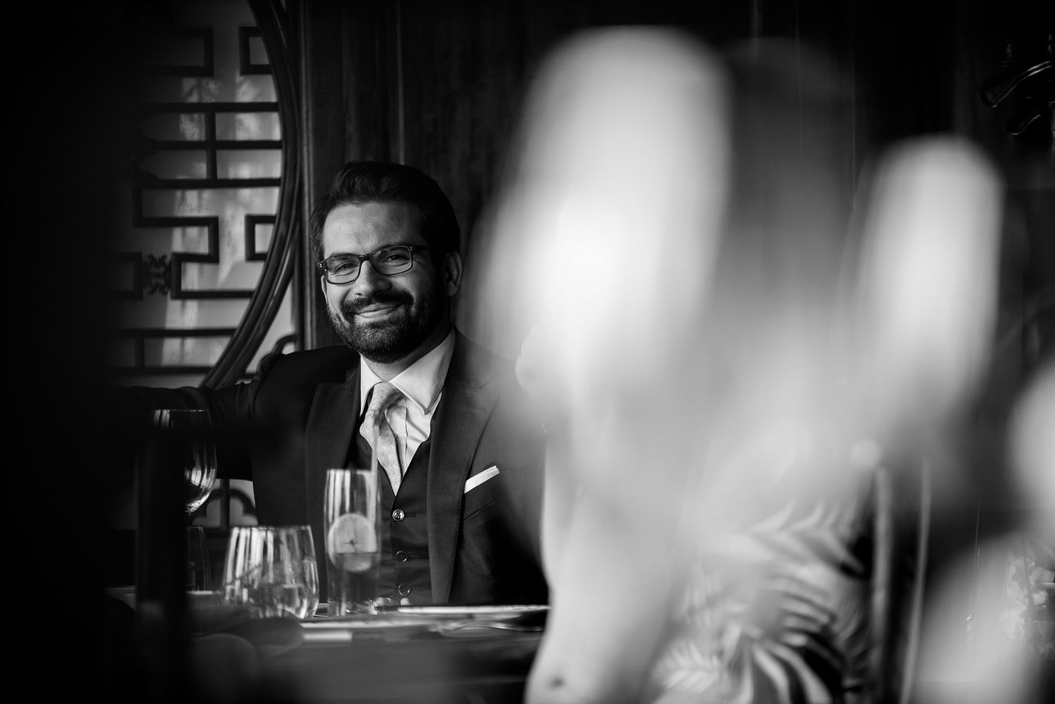Guests enjoying drinks and conversation around restaurant tables in dramatic black and white lighting.