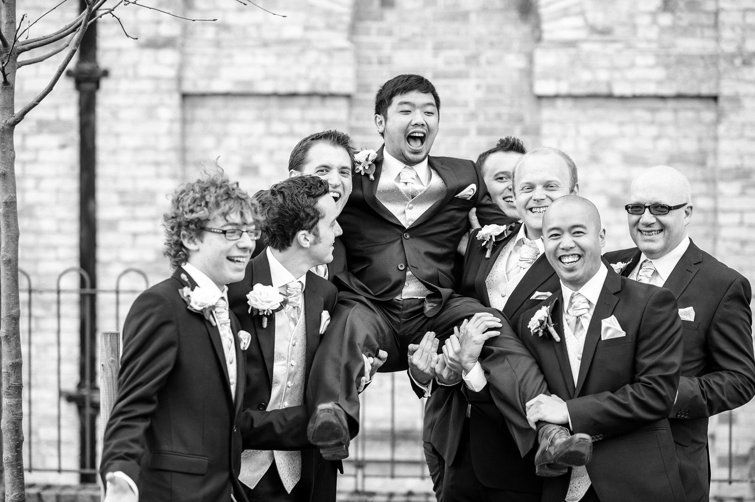 Groomsmen pose playfully while lifting someone in a black and white wedding photo.