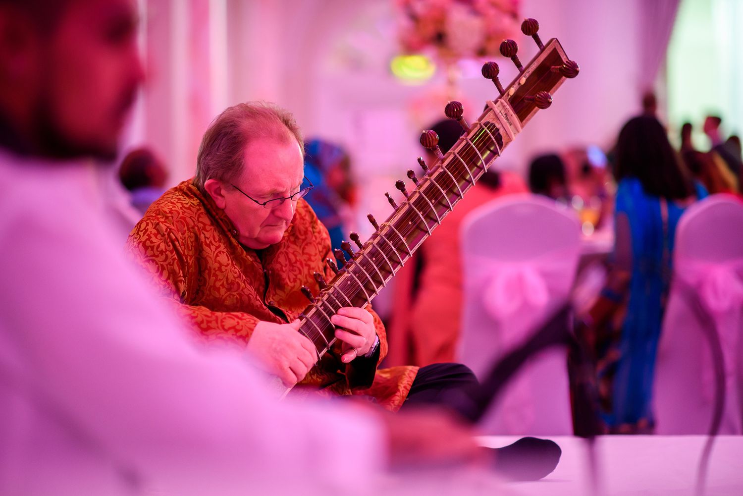 Musician playing sitar at cultural wedding celebration bathed in pink lighting.
