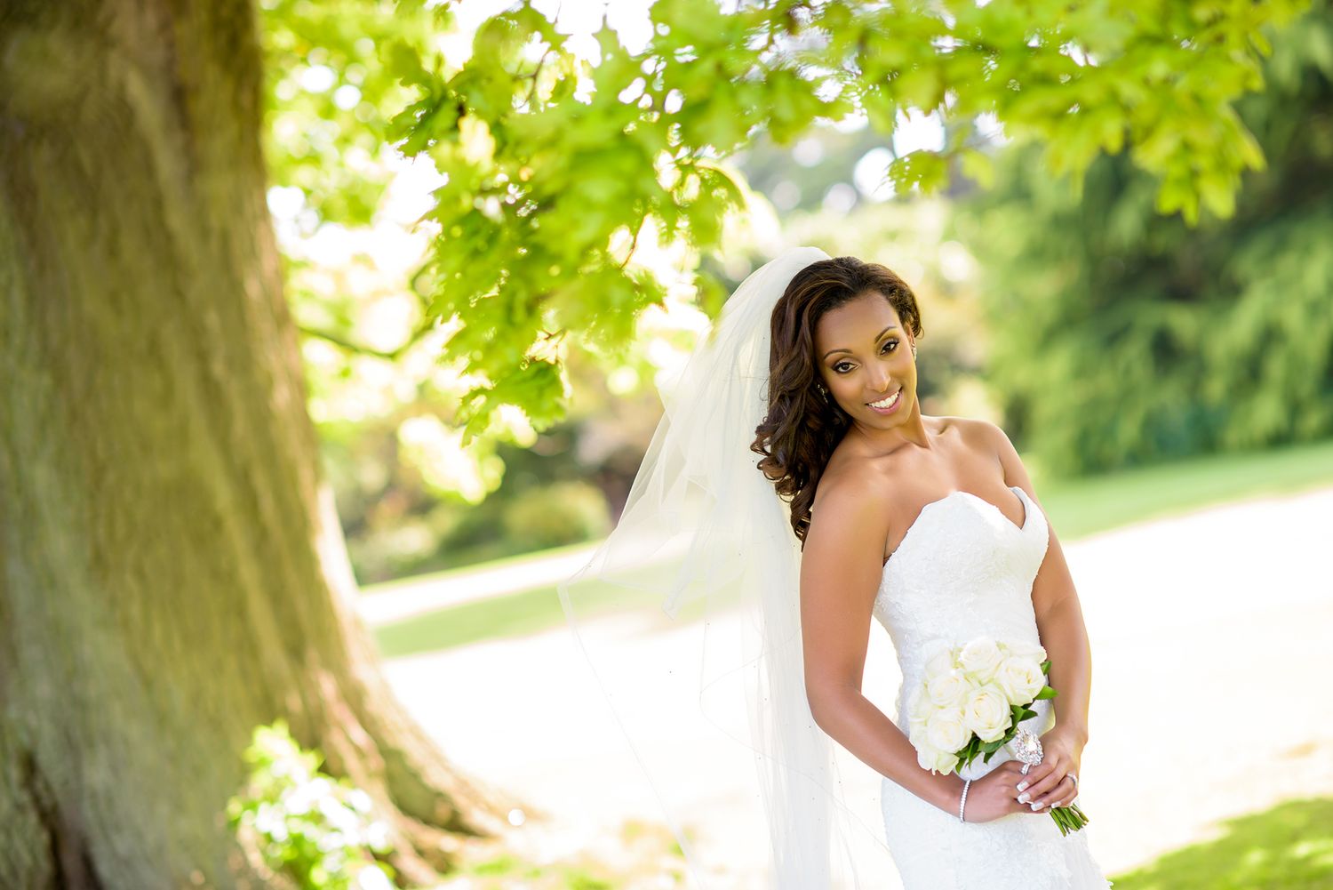 Bride in flowing white dress stands gracefully under lush summer foliage.