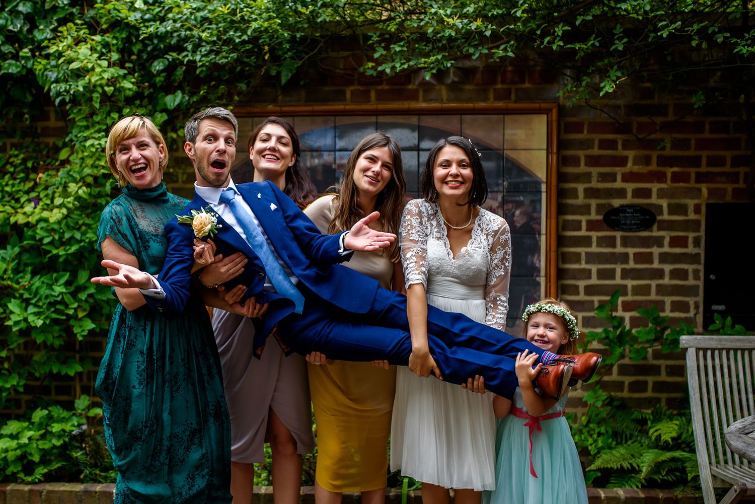 Wedding group poses playfully in front of brick building surrounded by greenery.