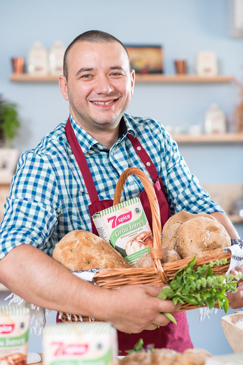 Baker in checkered shirt and red apron holding fresh bread baskets in bright kitchen.