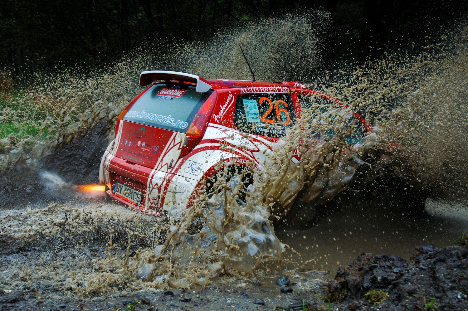 Red rally car splashing through muddy water during an off-road racing competition.