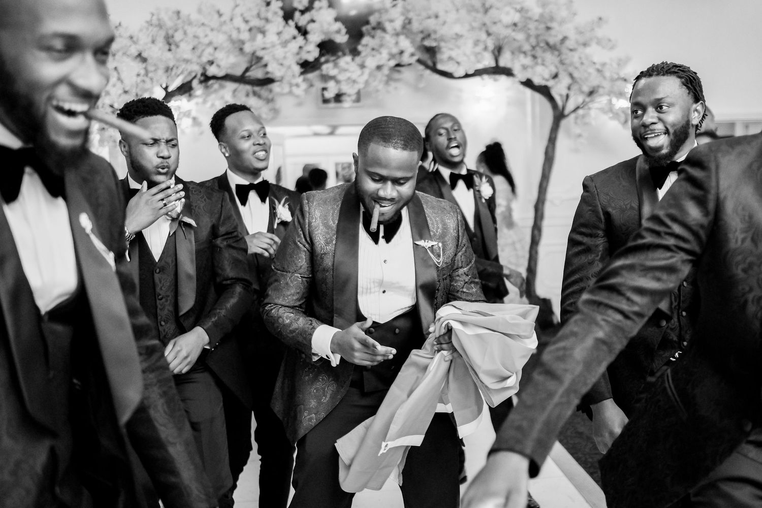 A black and white photo shows groomsmen sharing a lighthearted moment during wedding celebrations.