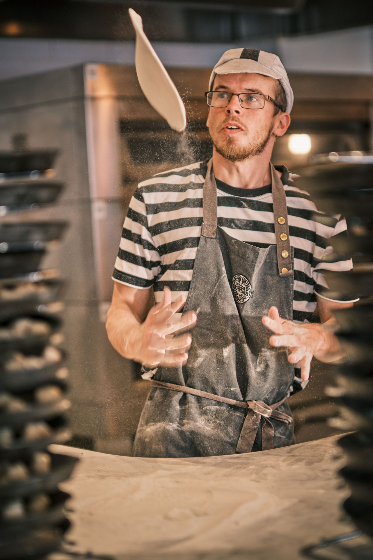 Pizza chef in striped shirt and apron tossing dough in a professional kitchen.