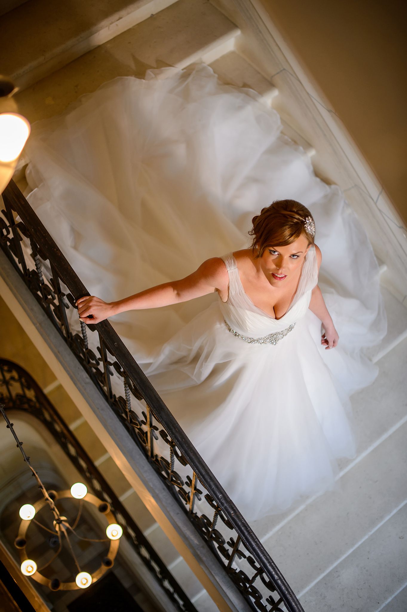 Bride in white wedding gown photographed from above on an elegant staircase with ornate railings.