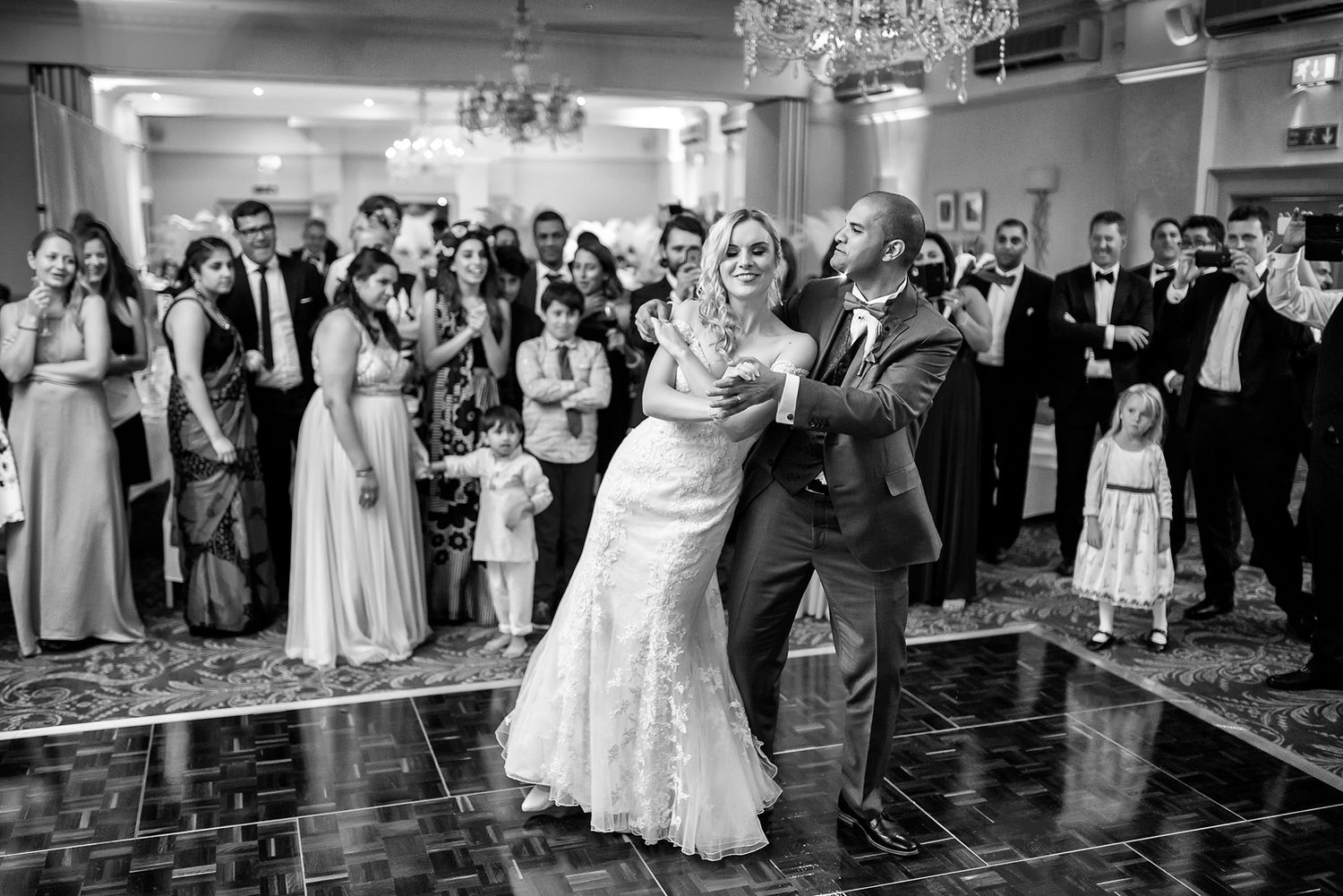 Black and white photo of wedding guests dancing and celebrating on a ballroom dance floor.