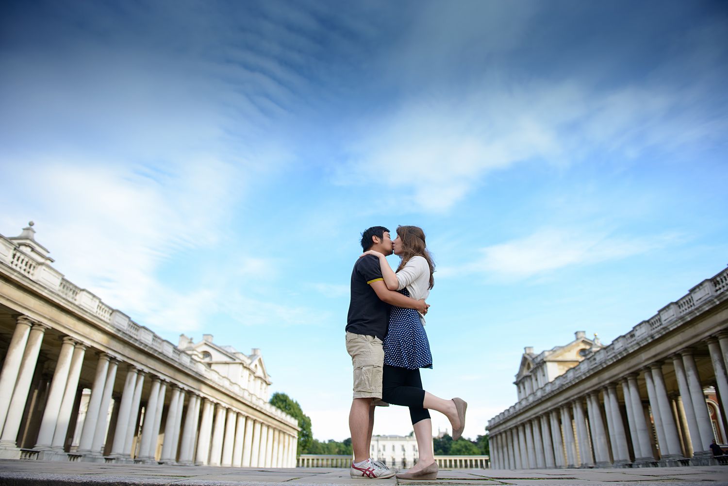 Couple embracing against a dramatic sky with classical architecture in the background.
