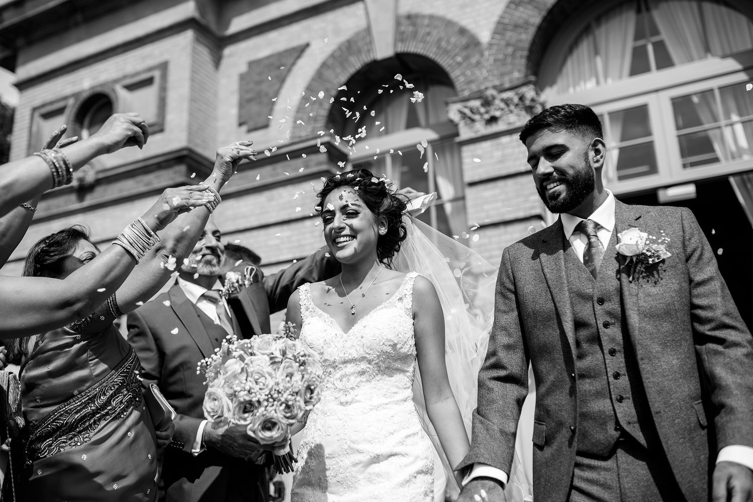 Black and white photo of wedding party celebrating with confetti outside historic building.