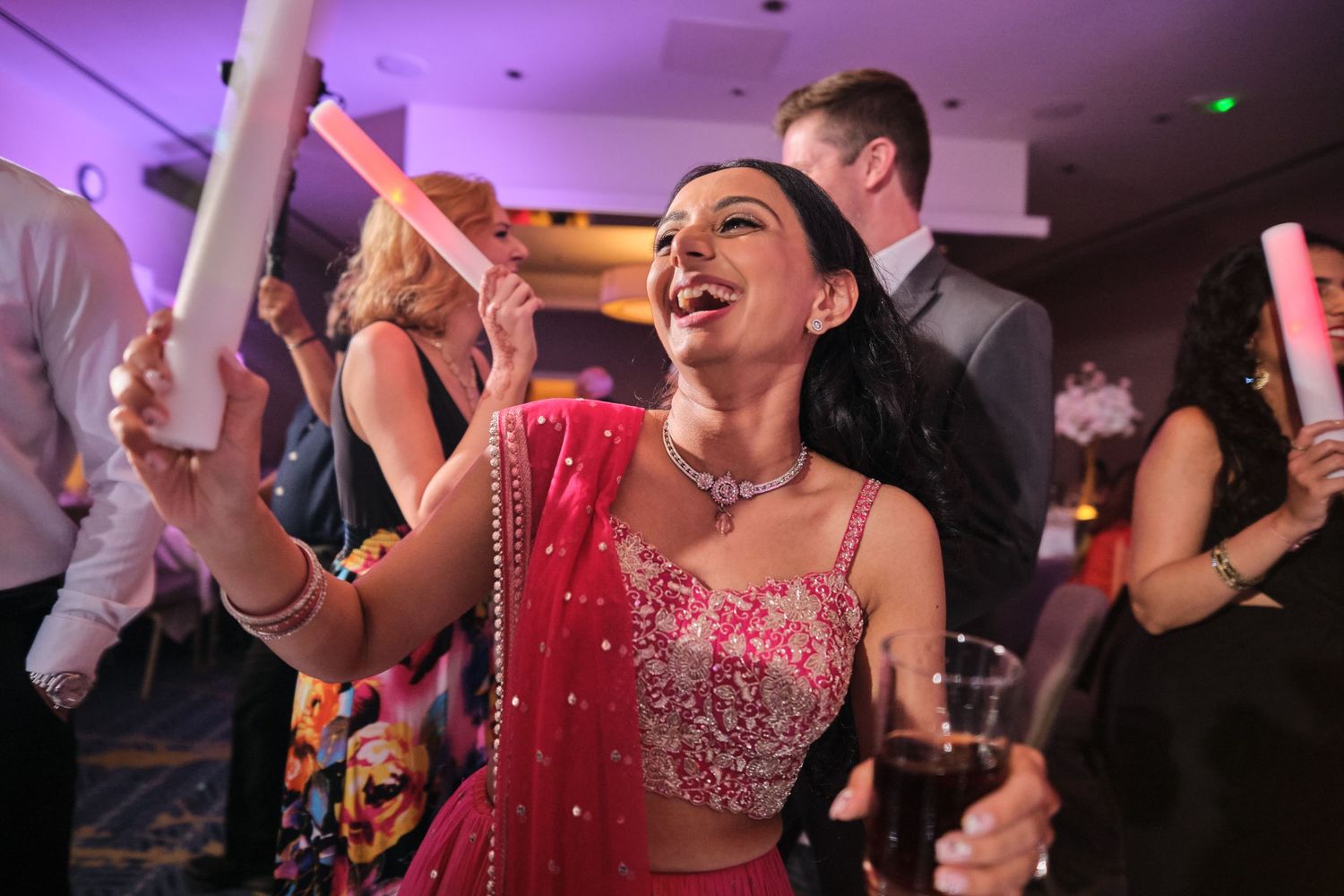 People dancing and celebrating at a wedding reception with colorful pink and purple lighting in the background.