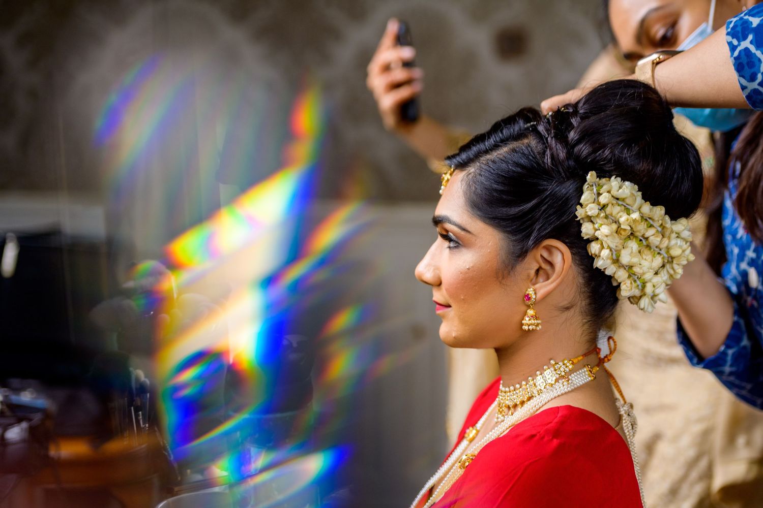 A person in a red sari with traditional jewelry and floral hair accessories during a wedding ceremony preparation.
