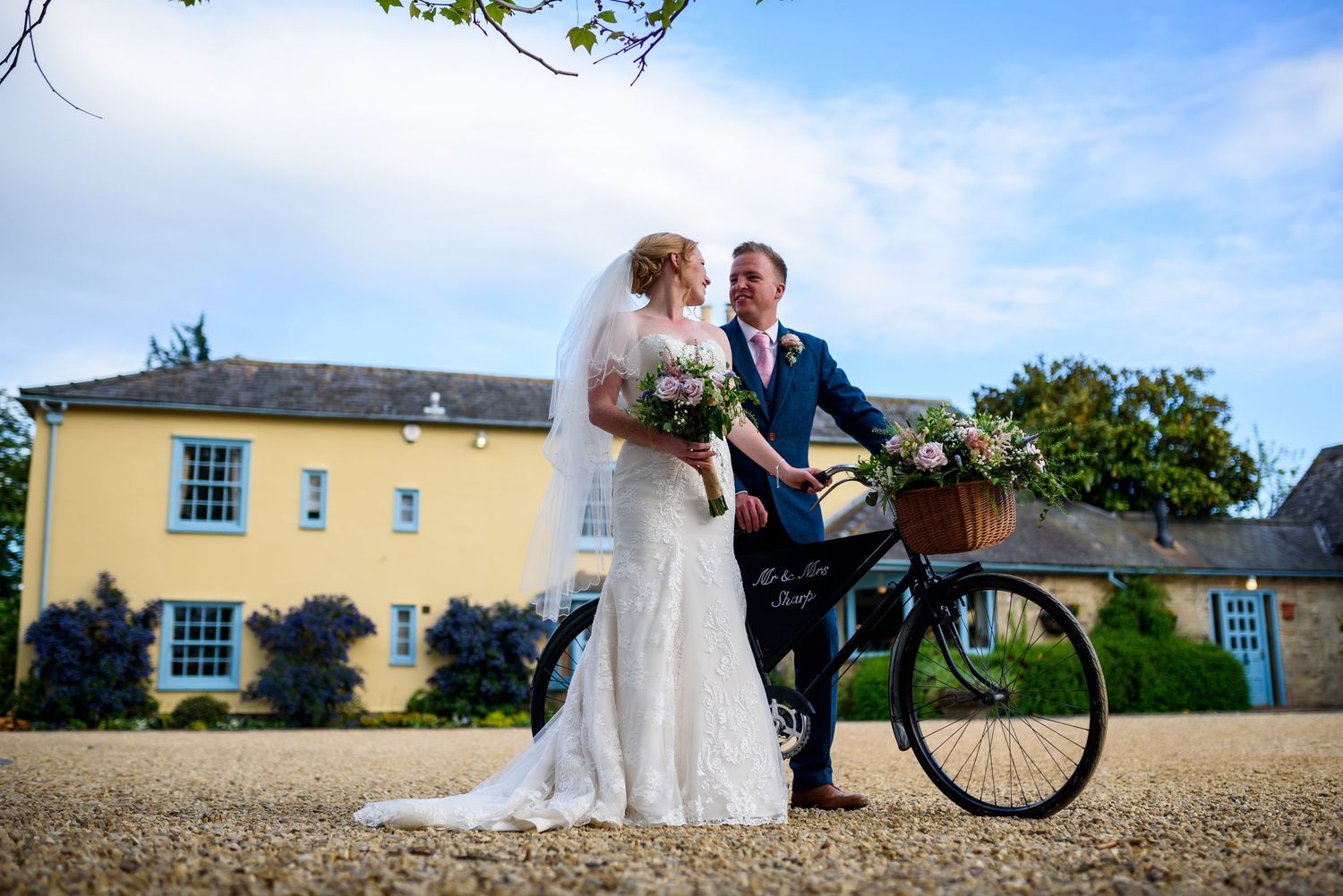 A couple poses with a vintage bicycle outside a yellow country house on their wedding day.