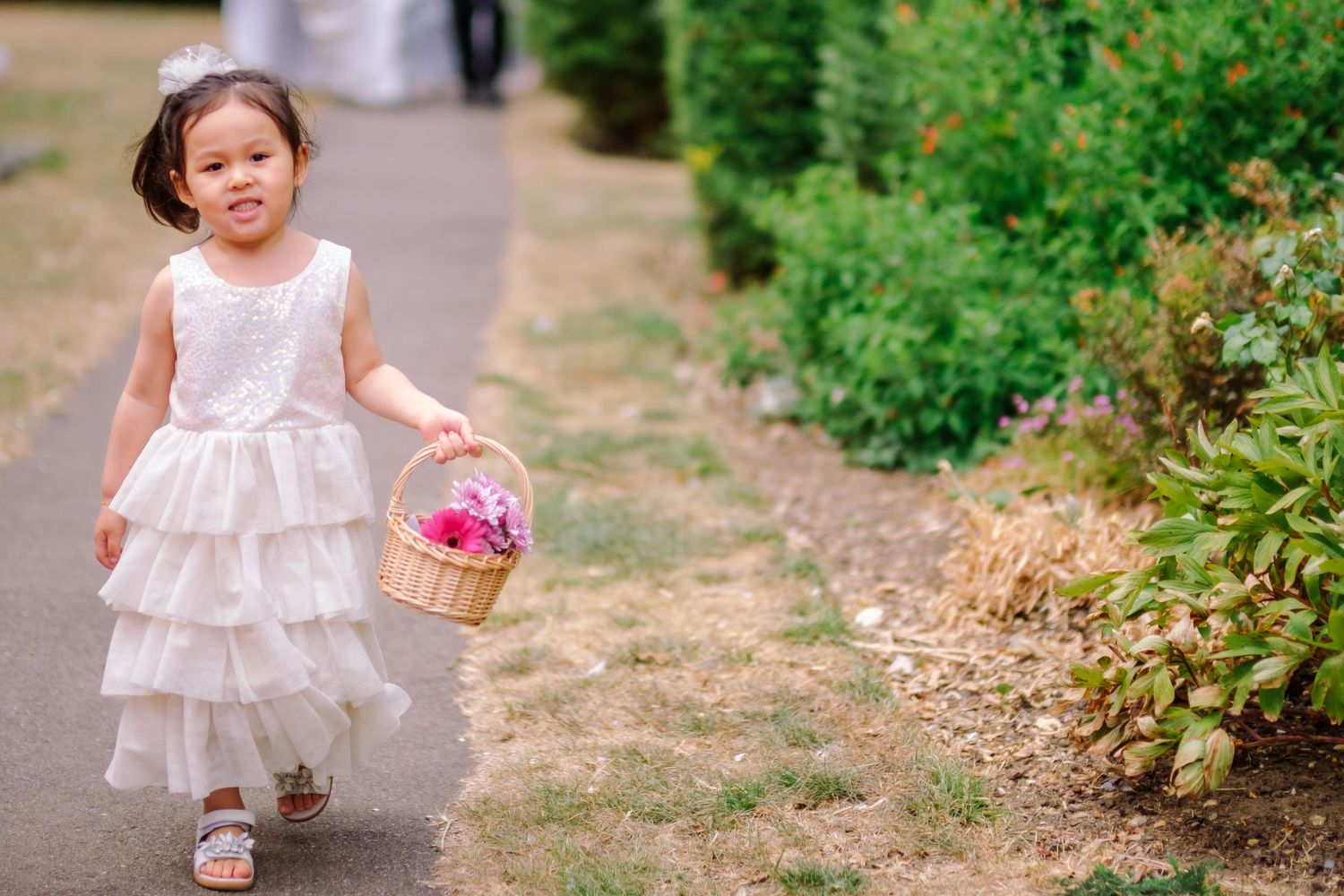 A small child in a white tiered dress carries a pink Easter basket while walking along a garden path.