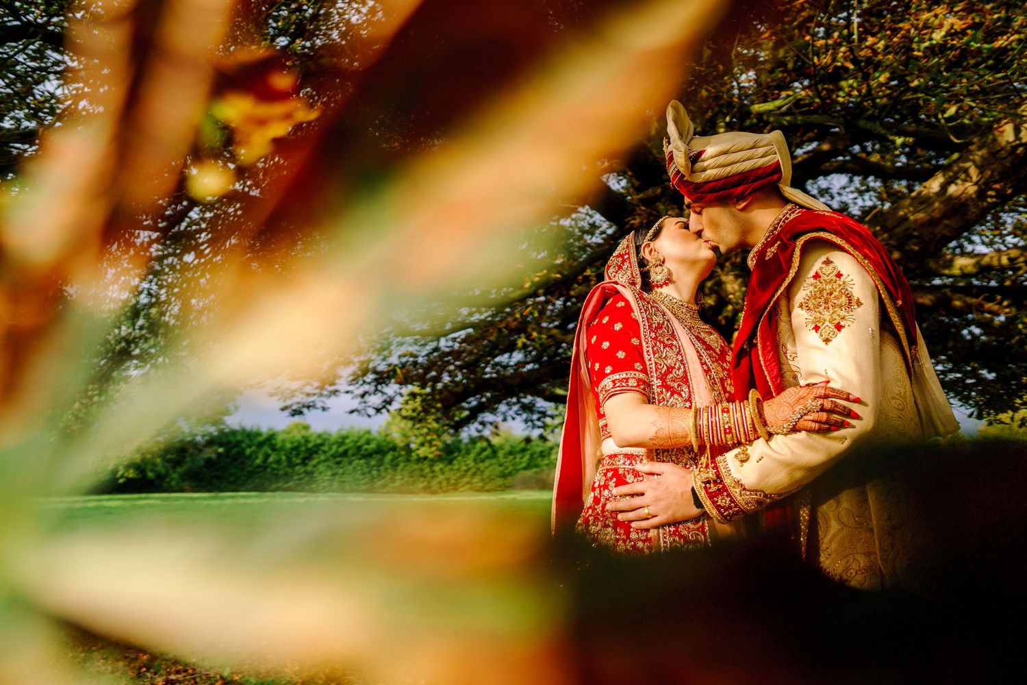 Romantic outdoor portrait through foliage featuring traditional Indian wedding attire.