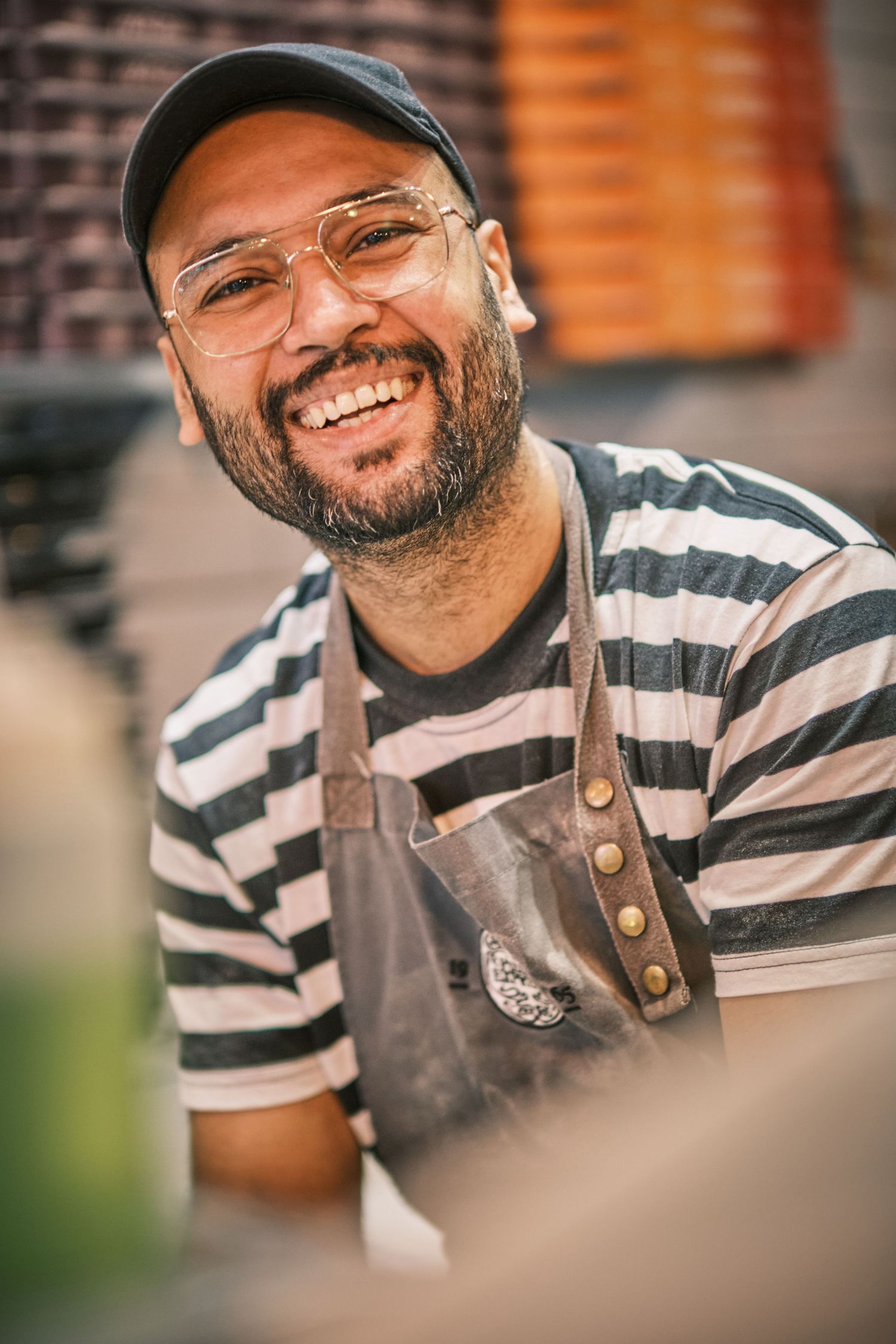 Chef wearing striped shirt and apron smiles warmly in restaurant kitchen setting.