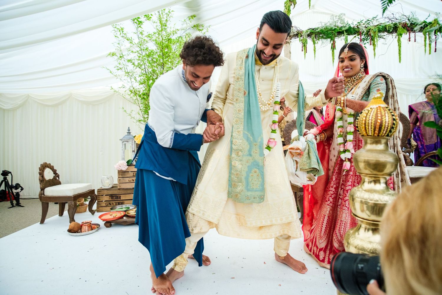 Wedding guests gather under a white tent decorated with floral arrangements for a traditional Indian ceremony.