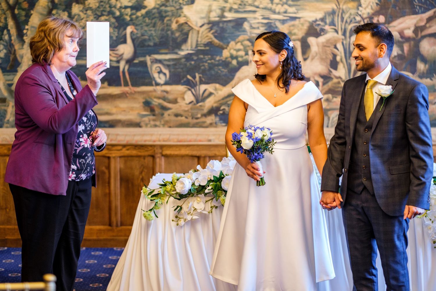 A wedding ceremony takes place in an ornate room with a tapestry backdrop and floral decorations.