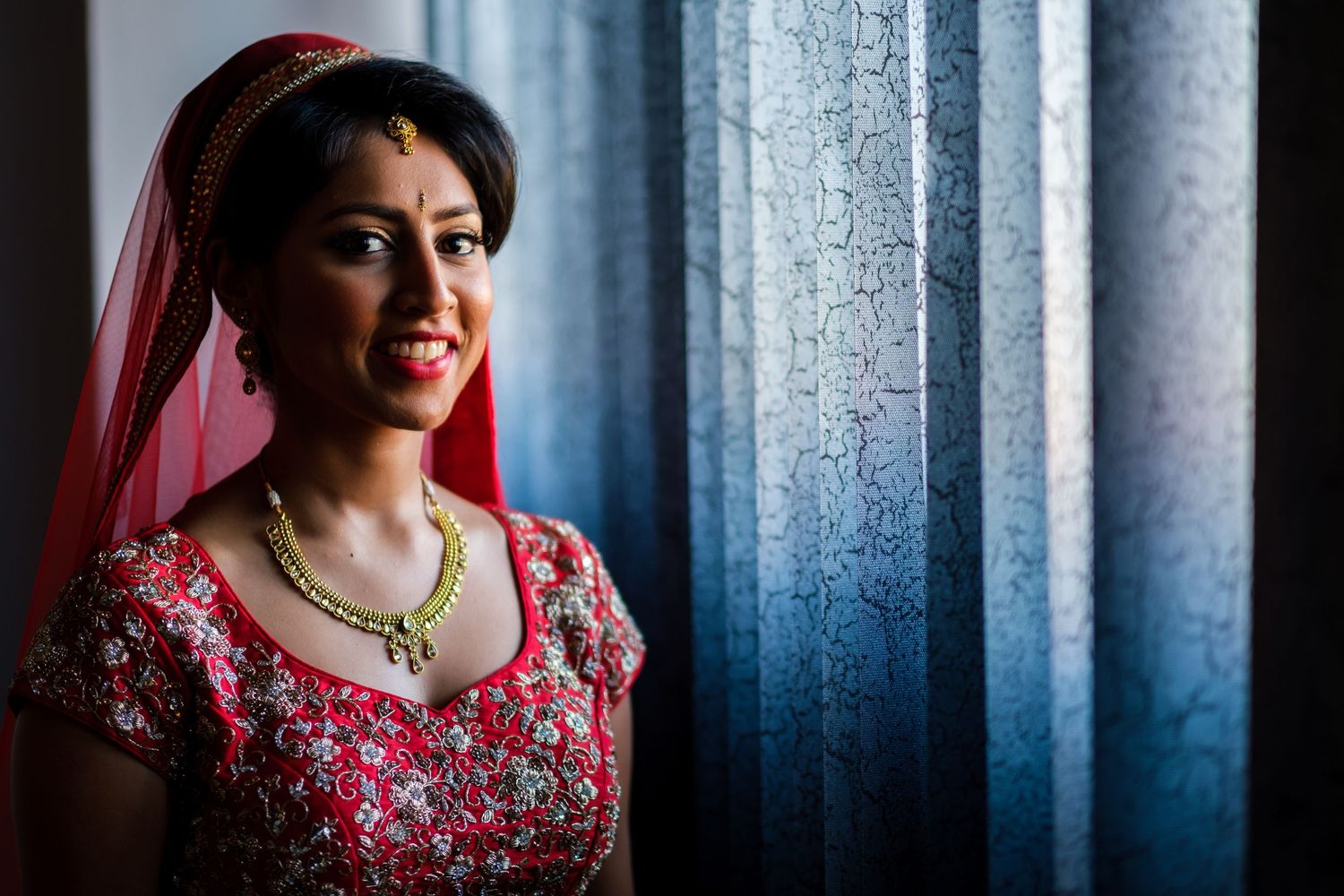 Traditional South Asian bride in red embroidered wedding attire poses by window with dramatic lighting.