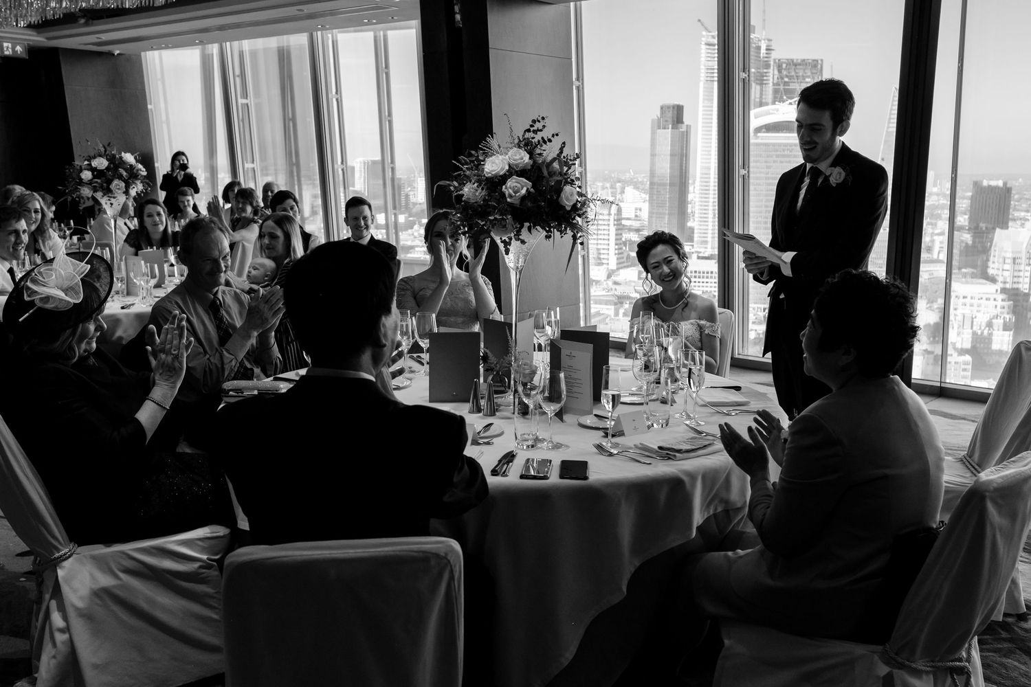 Silhouetted wedding reception guests enjoying dinner at tables by large windows.