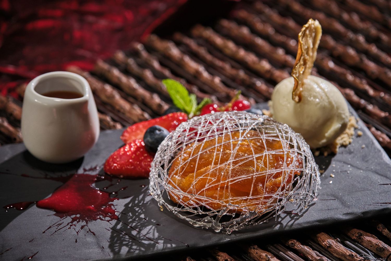 Elegant dessert with spun sugar nest, fresh strawberries, vanilla ice cream, and chocolate sauce on a dark slate plate.