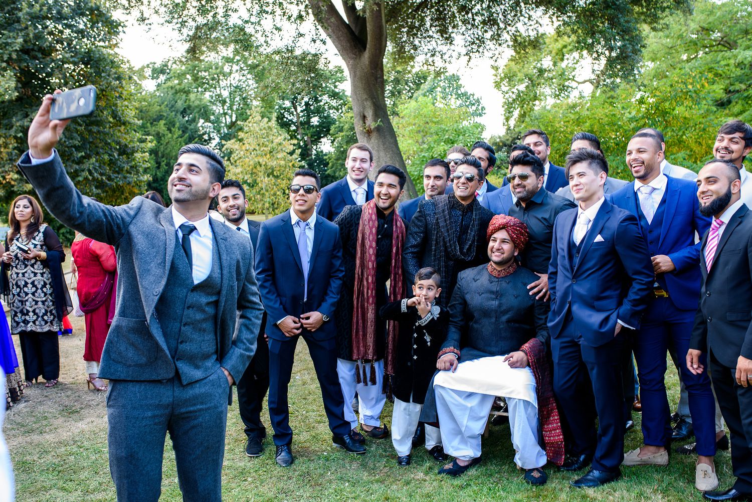 Large group of wedding attendees celebrating outdoors under tree canopy.