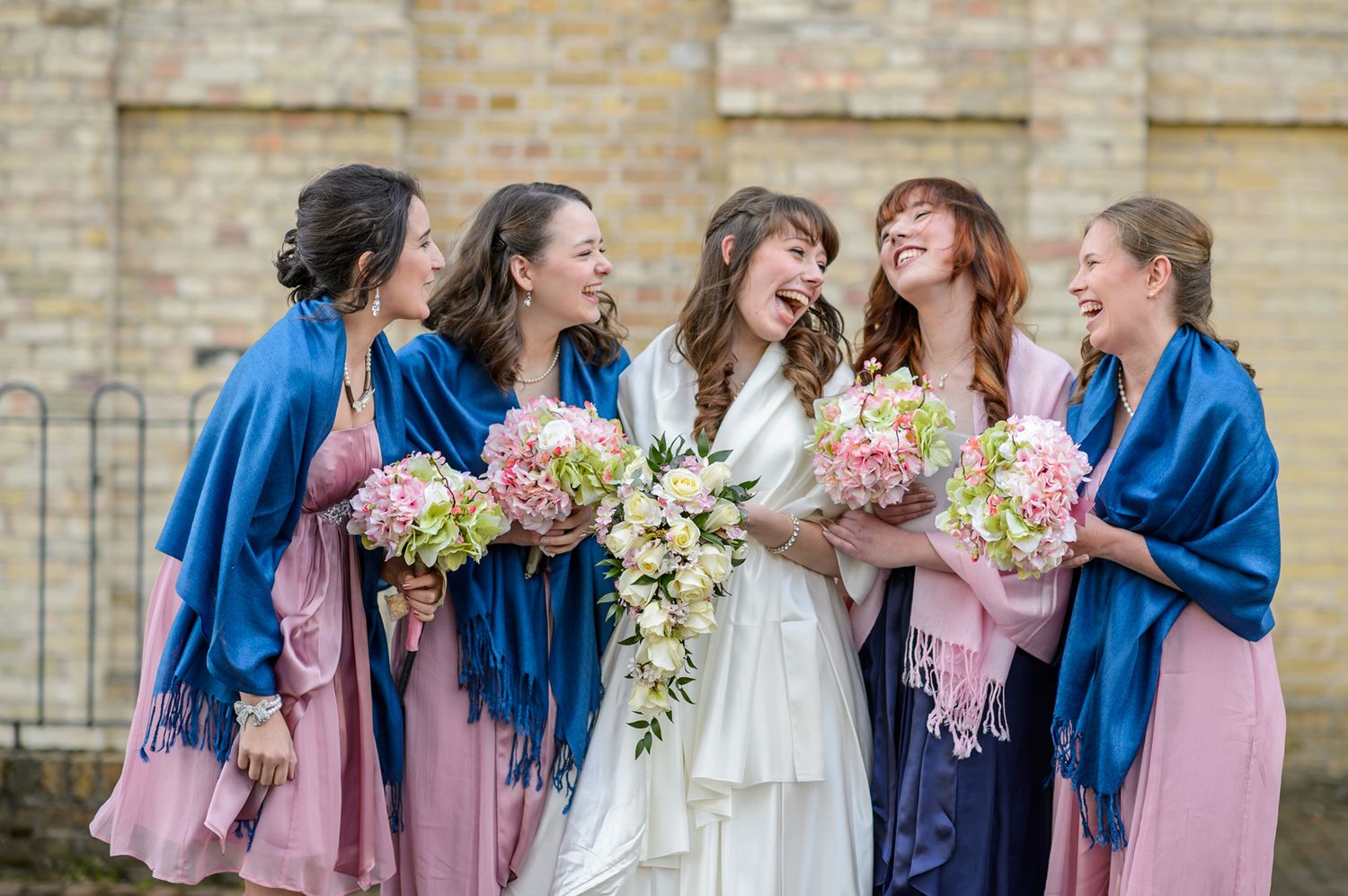 Bride in white with bridesmaids in blue shawls and pink dresses holding floral bouquets.