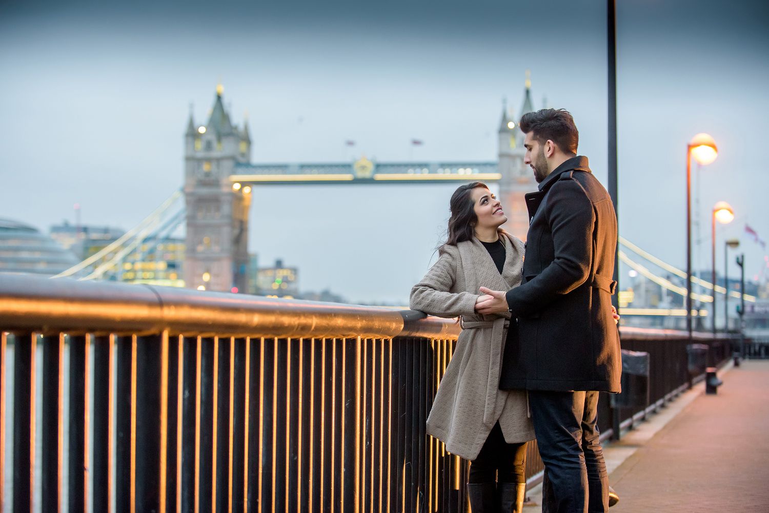 Silhouetted figures share intimate moment on bridge with urban skyline in background.