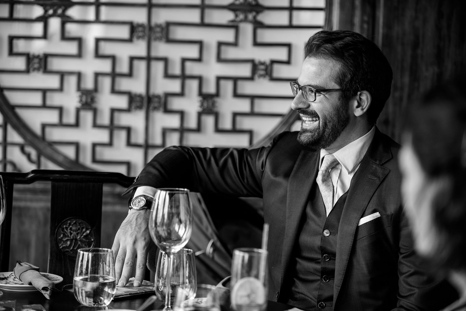 Black and white candid shot of people dining and conversing at an elegant restaurant with geometric wall patterns.