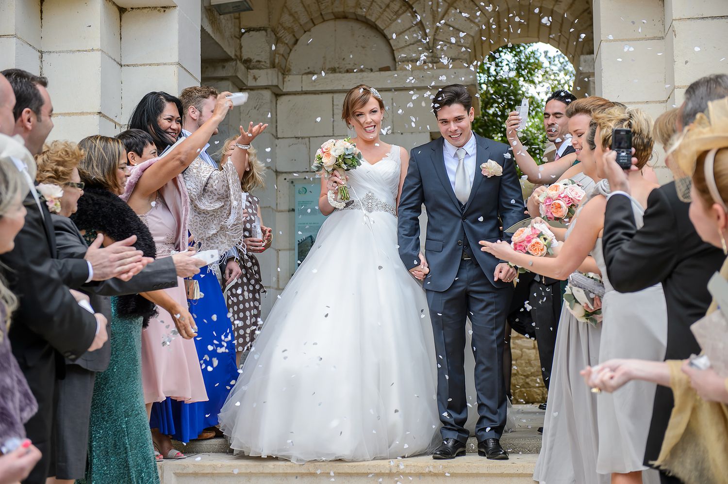 Wedding party celebrating with confetti toss outside historic stone building.
