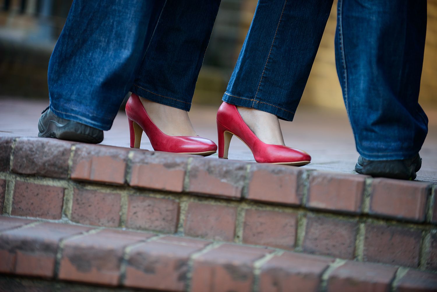 Close-up of red high heels and jeans near a brick wall outdoors.