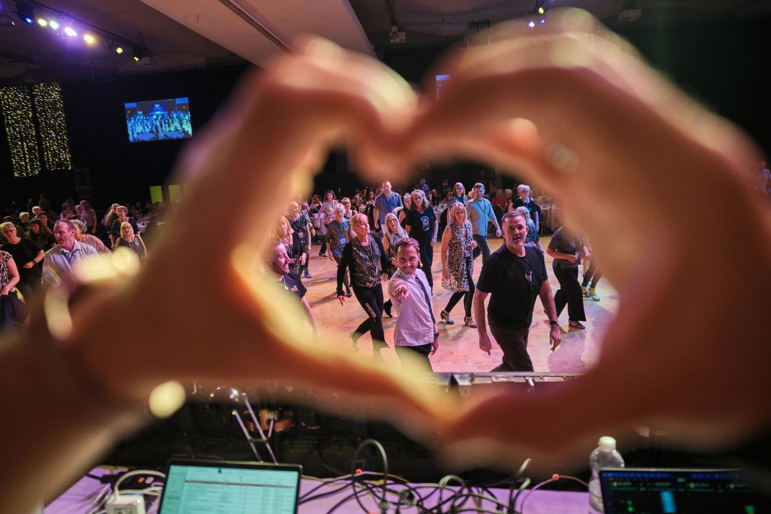 Dance floor crowd at a lively event with hands raised in the air creating heart shapes.