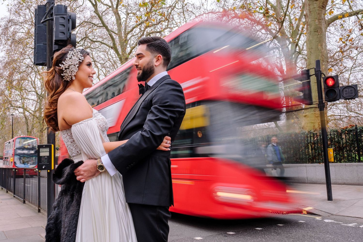 A couple embraces as a red London bus speeds past them on a tree-lined street during an autumn day.