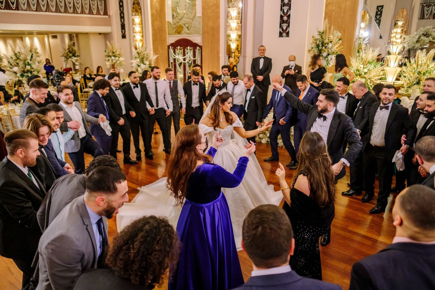 A group of guests gather in a circle around a woman in a royal blue dress at an elegant ballroom reception.