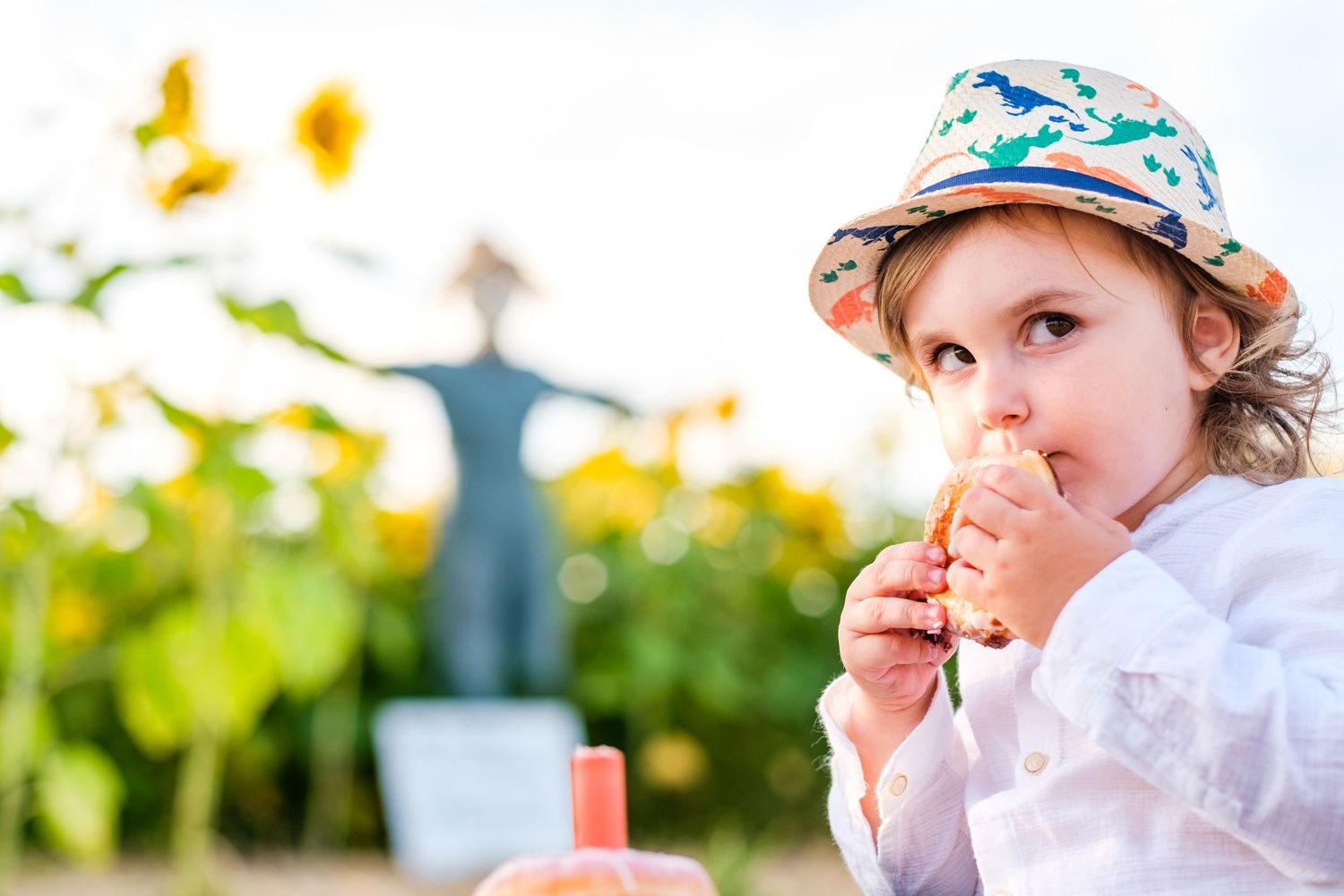 Young child in floral hat holds flower while exploring outdoors in bright sunlight.