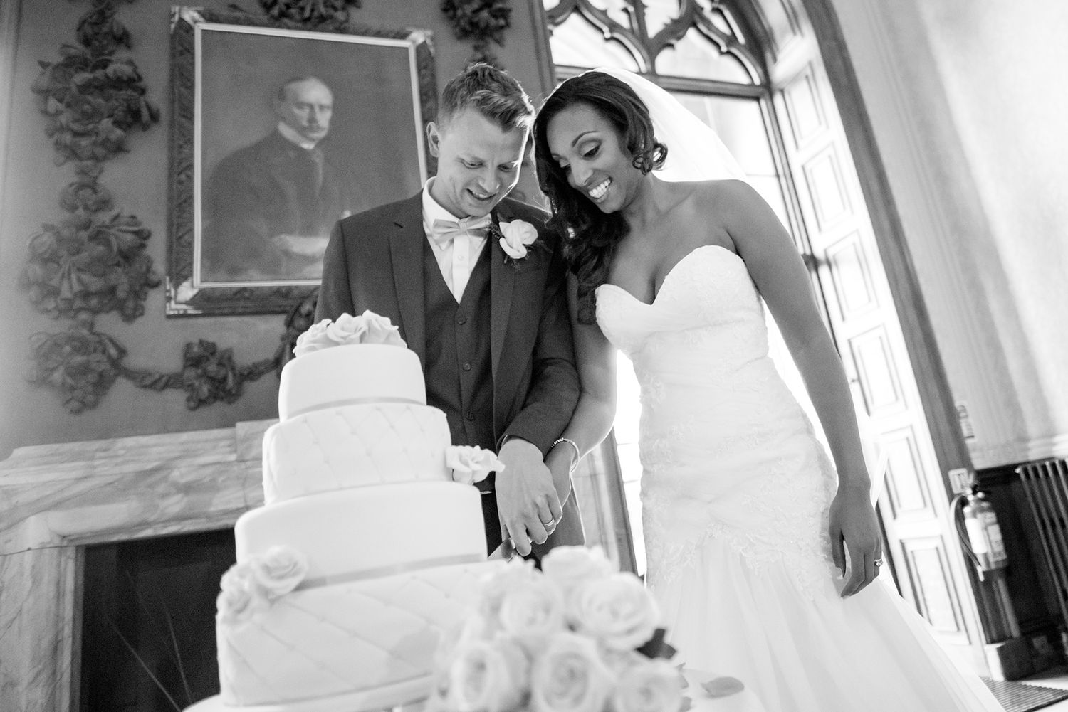 Newlyweds cutting their elegant white tiered wedding cake together in a ornate venue.