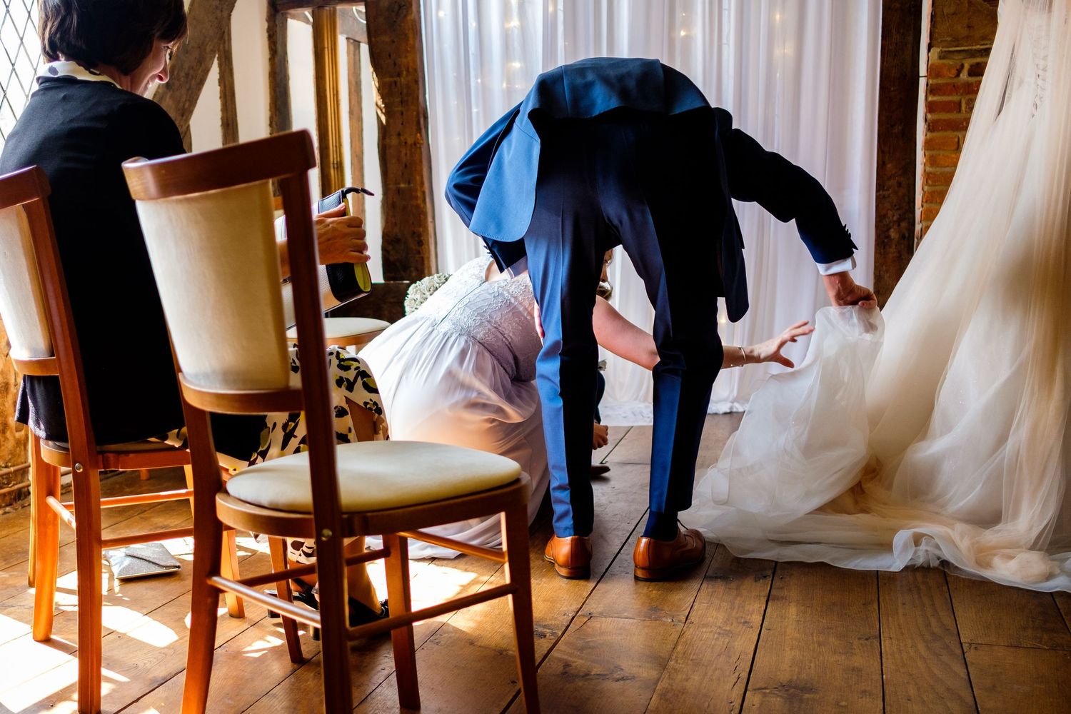 A candid wedding moment showing someone bending down to adjust a flowing white dress on a wooden floor.