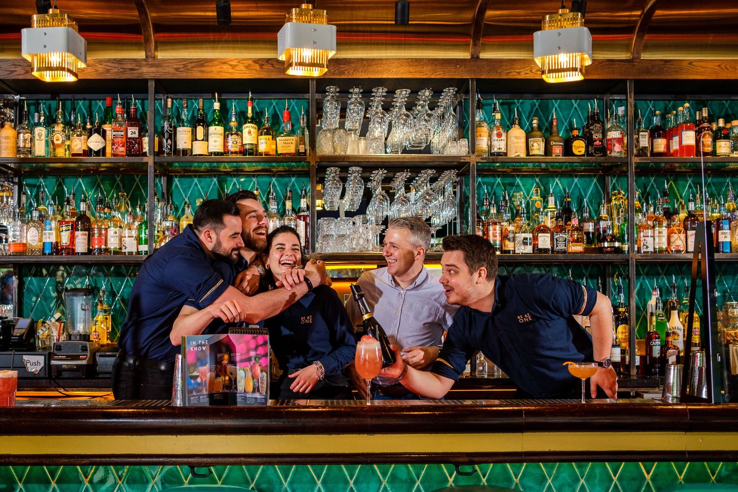 Bar staff posing together behind counter with extensive liquor bottle display.