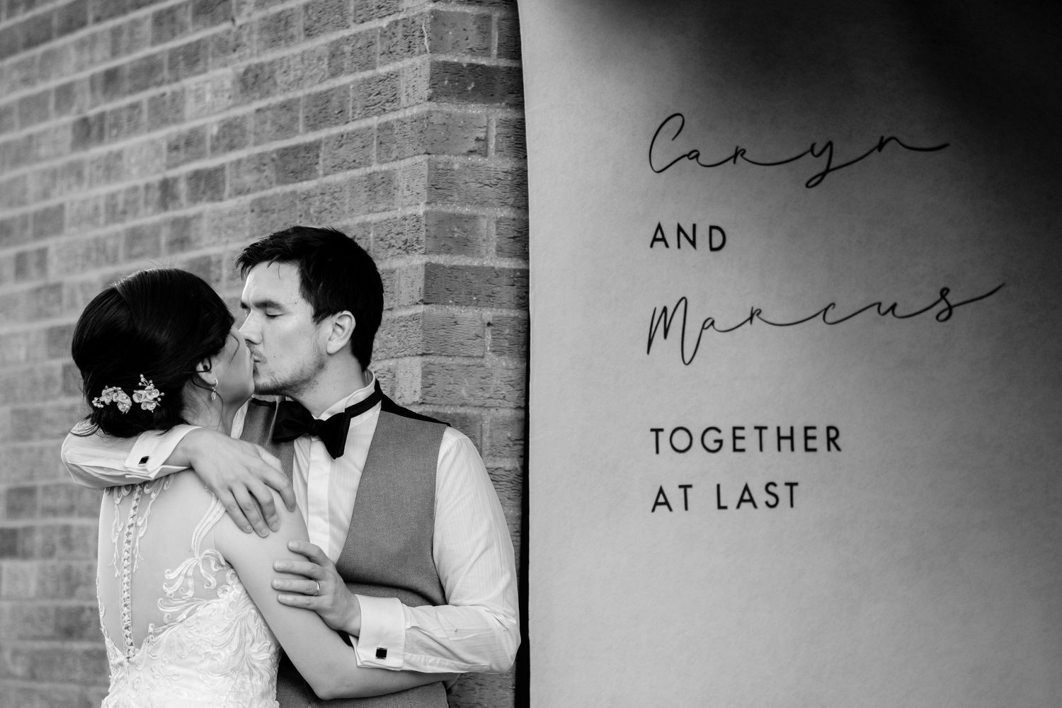 Black and white wedding photo with text reading 'Together at Last' on brick wall background.